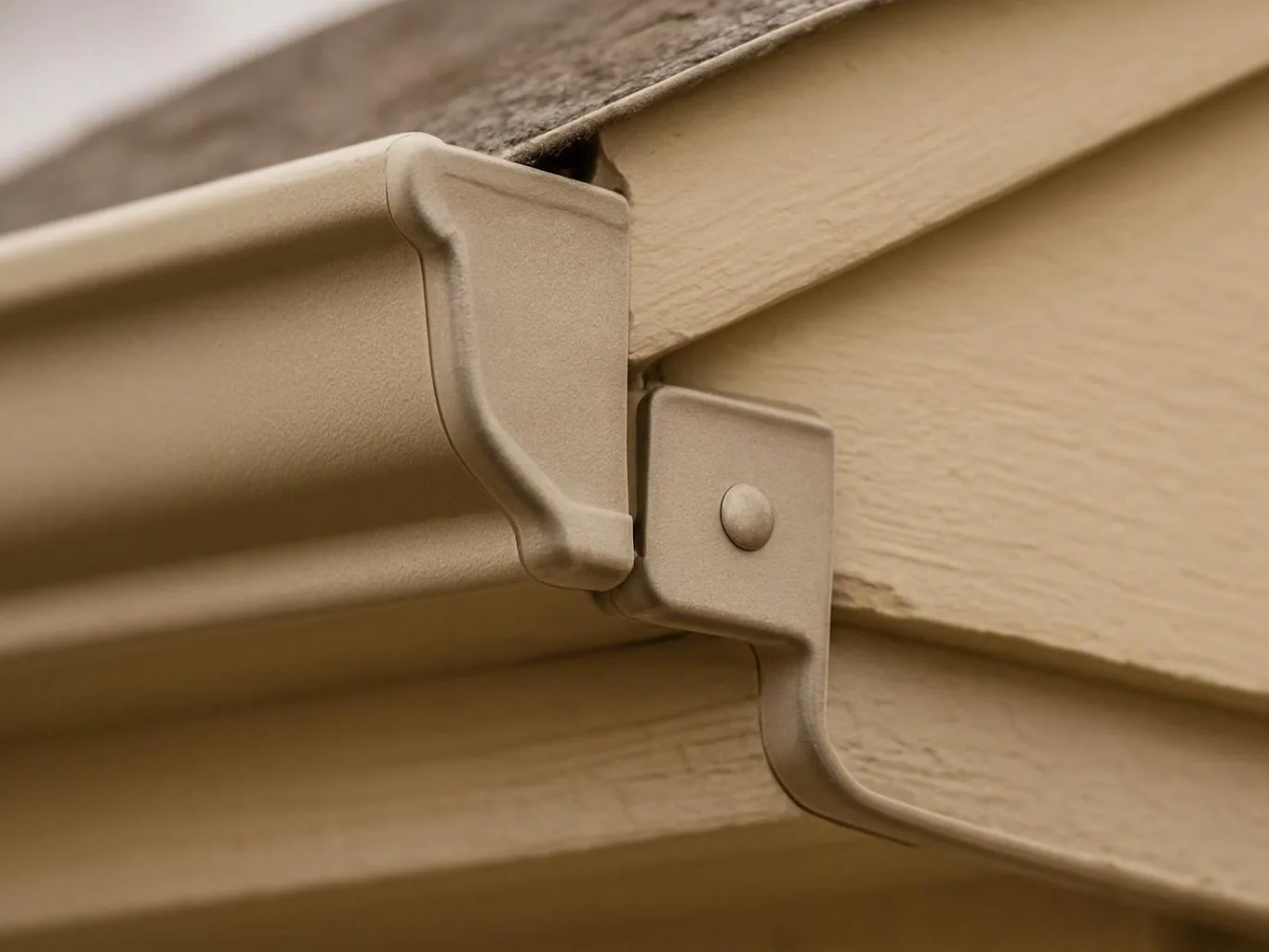 Close-up of a beige gutter bracket fastening under a shingled roof against wood siding