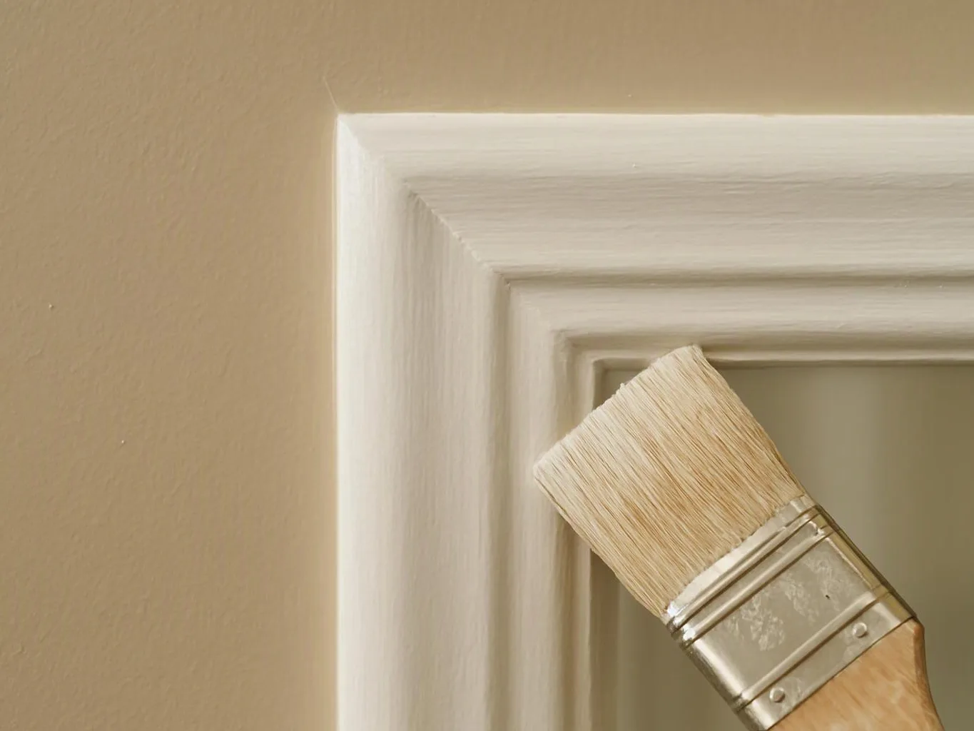 A paintbrush applies white paint to a door frame against a tan wall