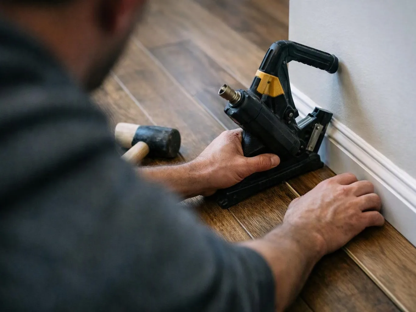 A person installs white baseboard trim with a black hand tool beside a rubber mallet on a wood floor