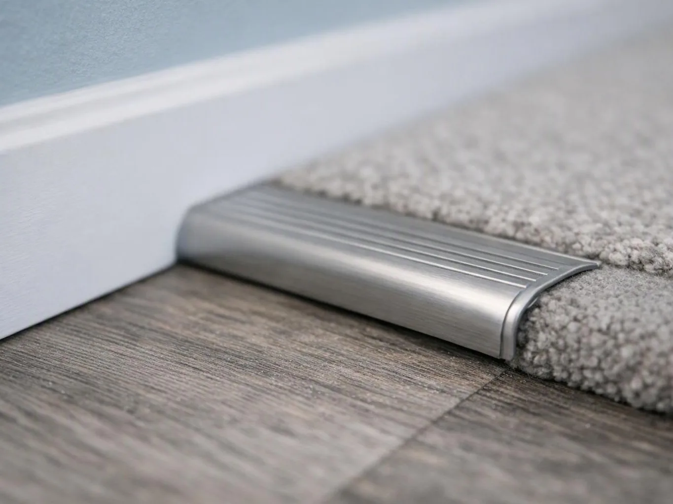 A silver floor transition strip joins wood-look flooring, carpet, and a white baseboard beside a blue wall
